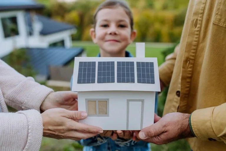 Hände halten ein Modellhaus mit Photovoltaikmodulen auf dem Dach
